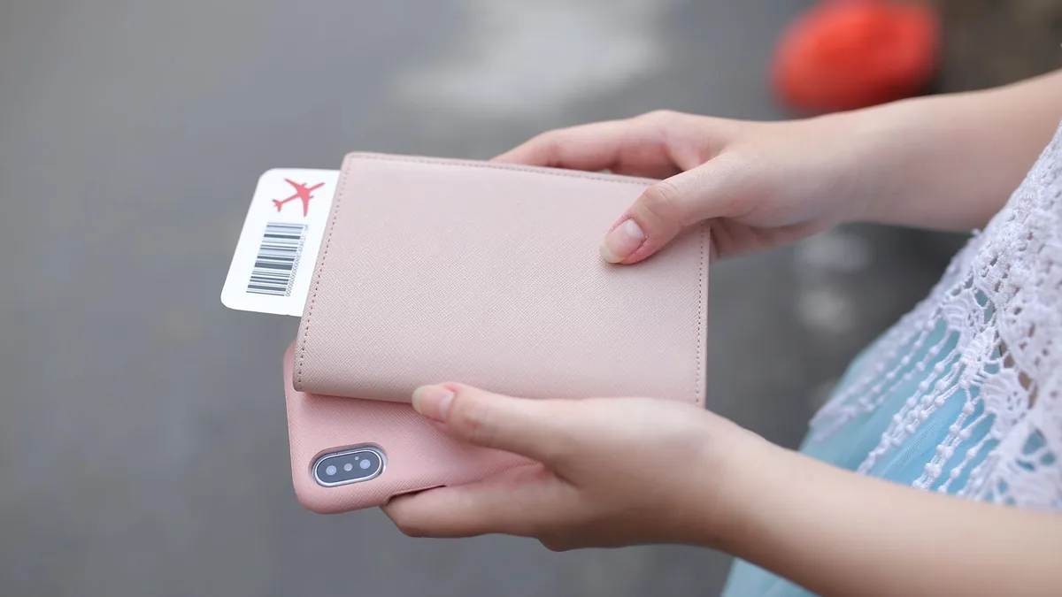 Hands holding a passport in a pale pink cover with a boarding pass tucked inside, a smartphone underneath