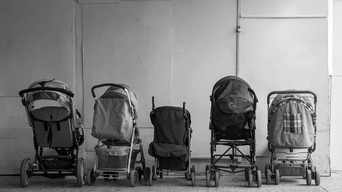Black and white photo of five folded-open pushchairs and prams parked in a row against a plain wall