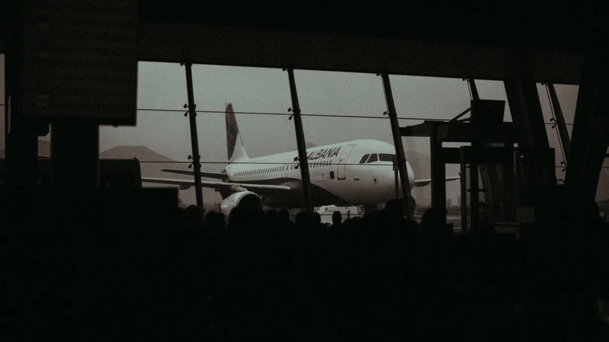 Silhouetted passengers waiting in a dim airport terminal, a parked airliner visible through the tall windows