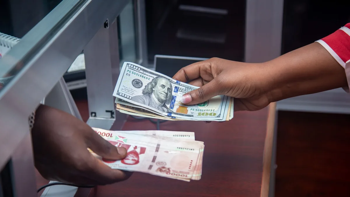 Hands passing US dollar notes and other banknotes across a glass-screened currency exchange counter