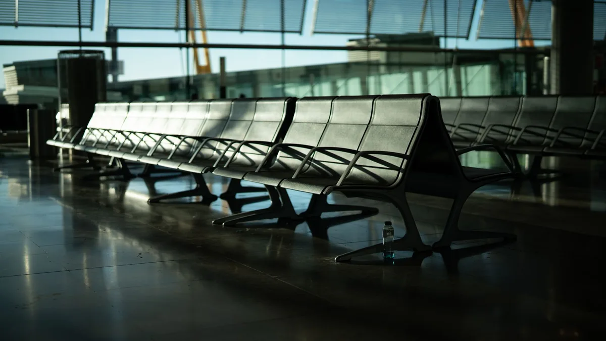 Rows of empty metal seats in a quiet airport terminal waiting area, sunlight through tall glass windows