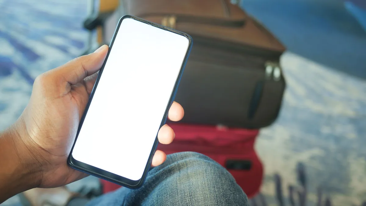 A traveller's hand holding a smartphone with a blank screen, with a suitcase and bag in the background