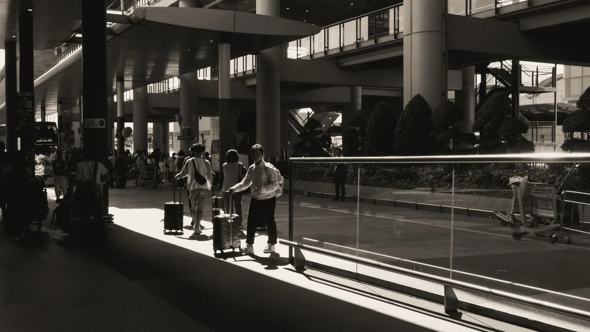 Black and white view of travellers with suitcases walking through a sunlit modern airport terminal concourse