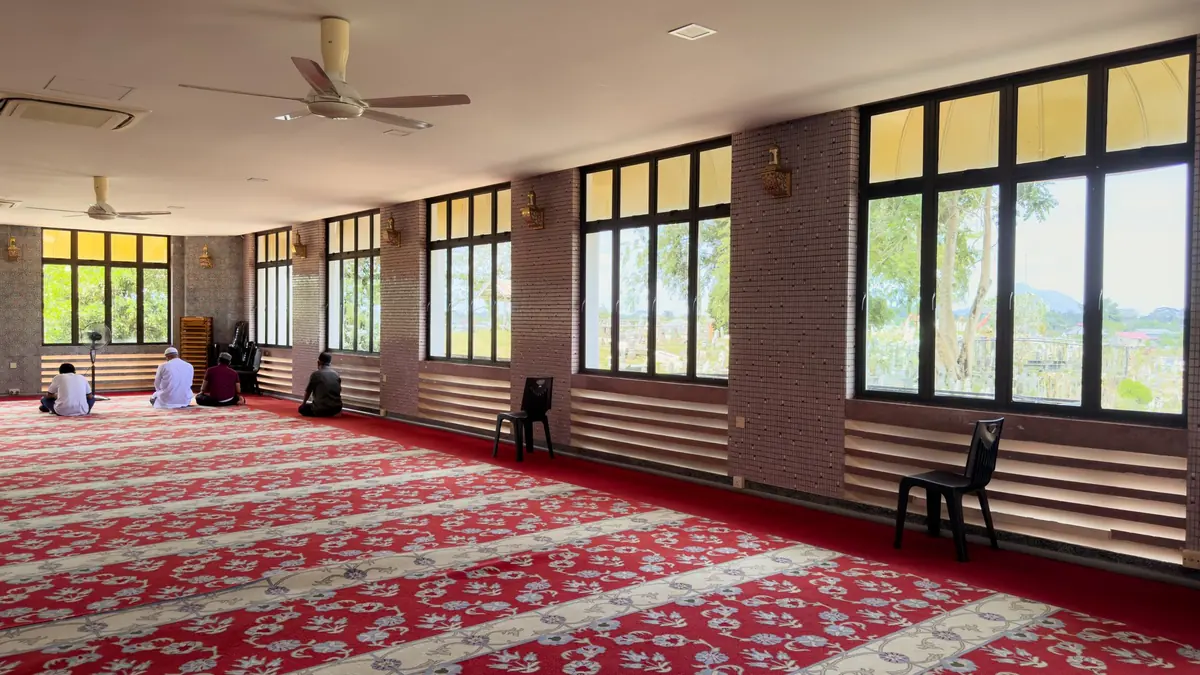 Quiet prayer room with a red patterned carpet, ceiling fans and a row of windows, a few worshippers seated far off