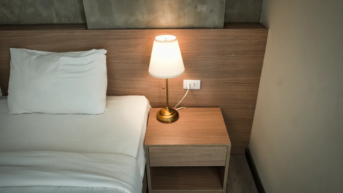 Simple hotel room with a made single bed, white pillow and a lit lamp on a wooden bedside table