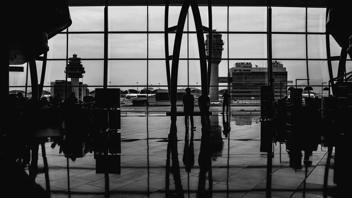 Black and white view of travellers silhouetted against tall airport terminal windows overlooking the apron