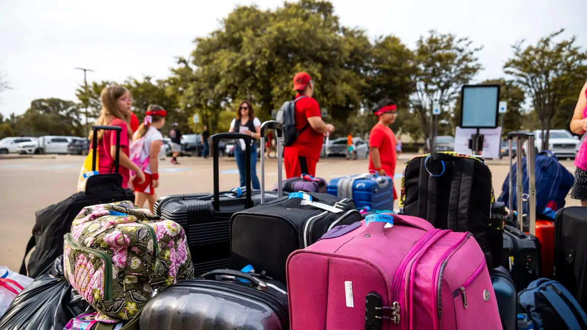 A large pile of colourful suitcases and bags grouped together outdoors, with travellers standing behind them
