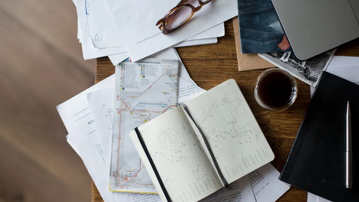 Overhead view of a wooden desk with an open notebook, folded maps, papers, a laptop, glasses and coffee for trip planning