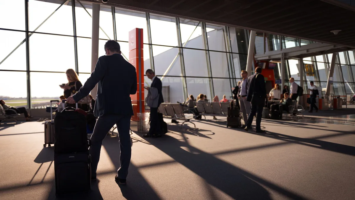 Travellers with wheeled carry-on luggage walking through a sunlit glass-walled airport terminal