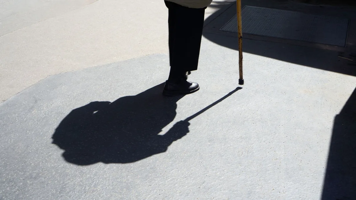 Older person walking with a wooden cane on sunlit pavement, face out of frame, a long shadow cast beside them