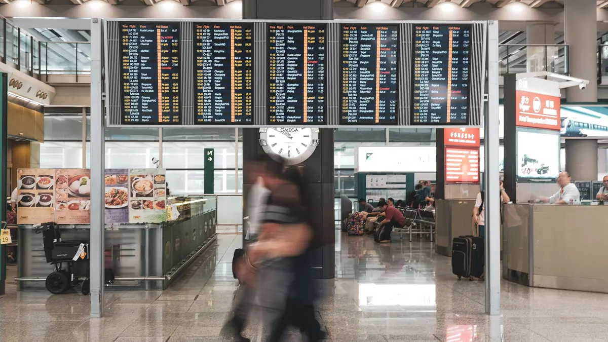 Travellers blurred in motion walking beneath a bank of flight information screens in a bright airport terminal