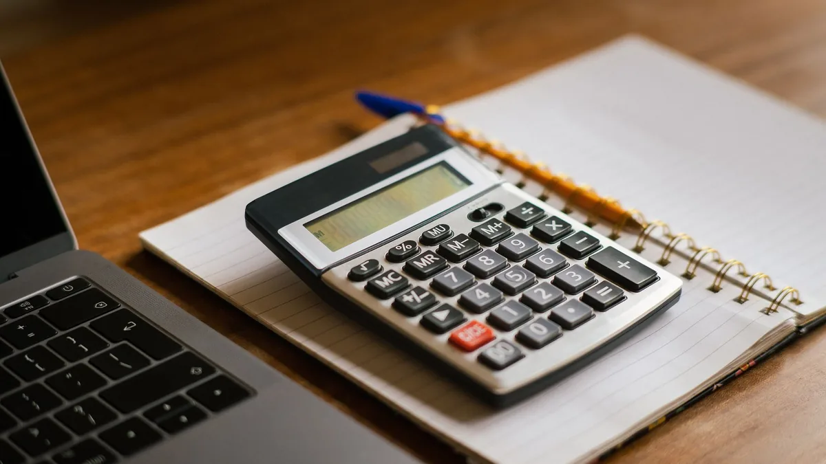 Calculator resting on an open spiral notebook beside a pen and laptop on a wooden desk, ready for budgeting