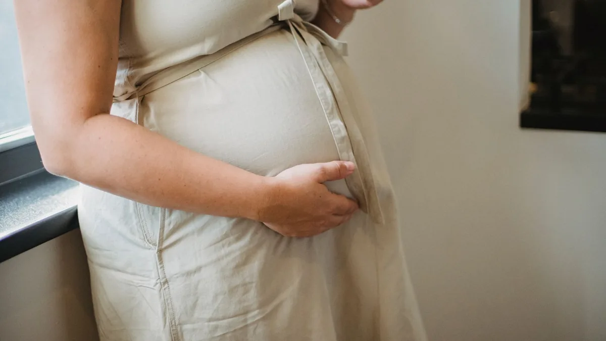 Pregnant woman in a beige dress, face out of frame, resting a hand on her bump while checking her phone by a window