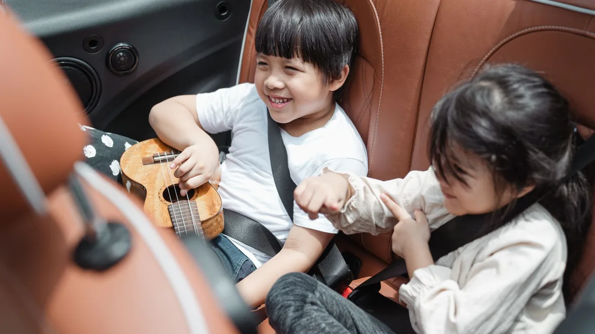 Two young children smiling in the back seat of a car with seat belts fastened, one holding a small ukulele