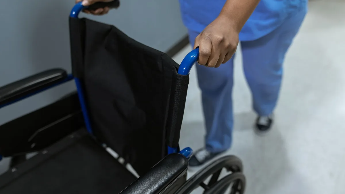Close-up of a staff member in blue scrubs pushing an empty black wheelchair along a hospital corridor floor