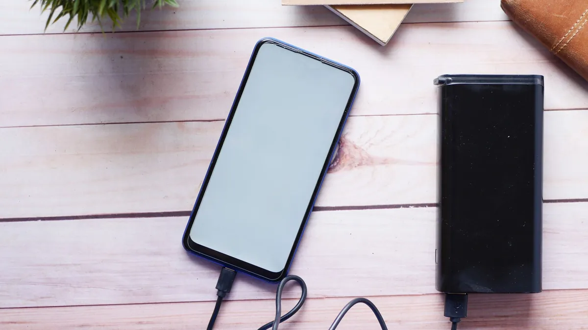 Smartphone with a blank screen connected by cable to a black power bank on a light wooden desk