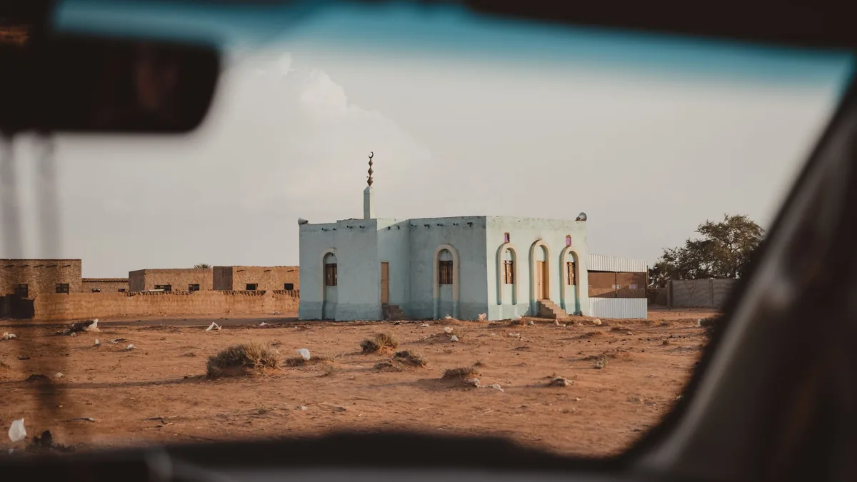 A small pale blue roadside mosque with a minaret seen through a car window across dry, open ground