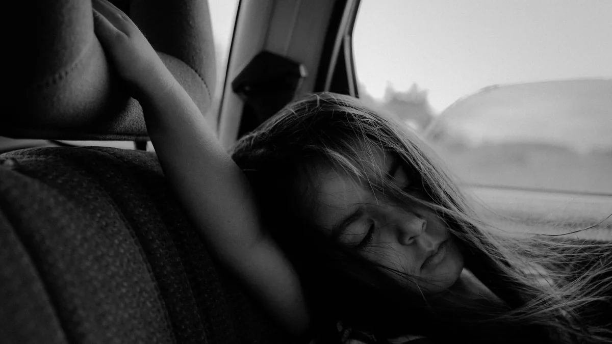 Black and white photo of a young girl asleep against the back seat of a car, hair across her face, with open countryside outside the window