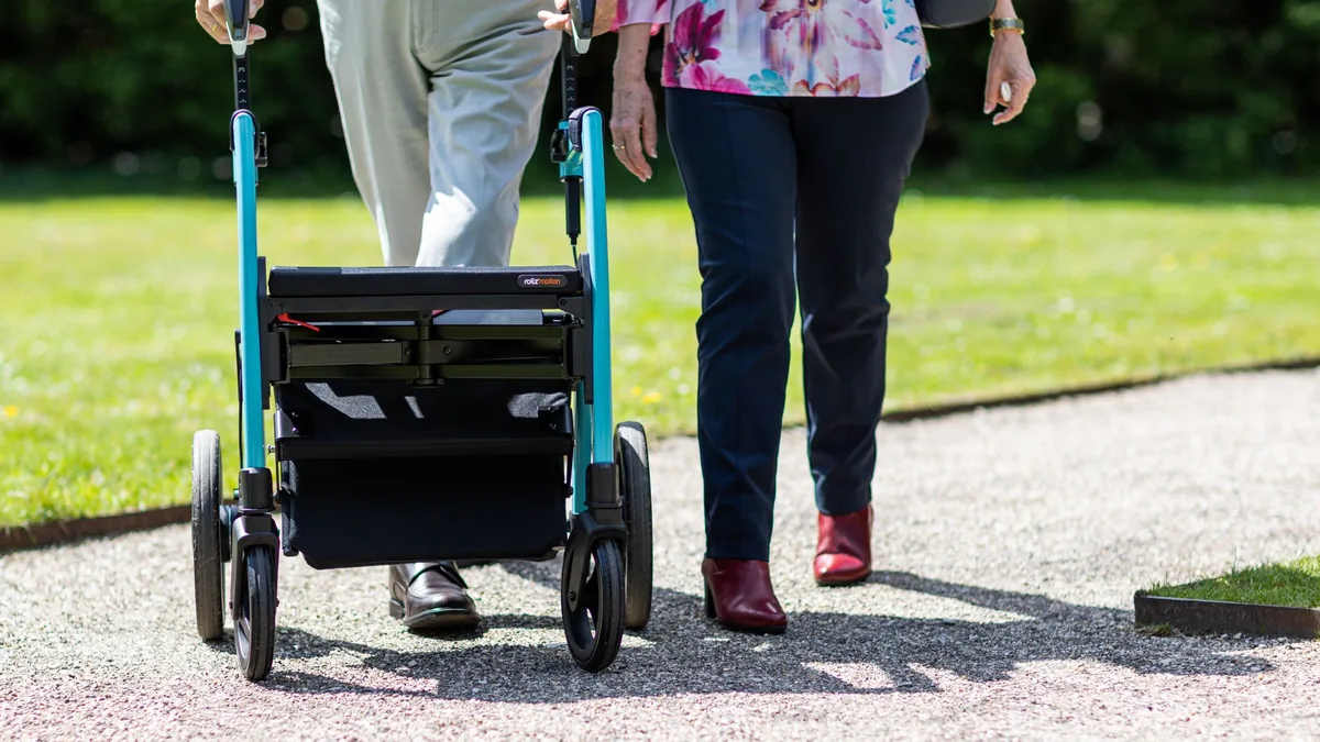 An older person pushes a wheeled rollator walker along a gravel path beside a companion, with a lawn behind them