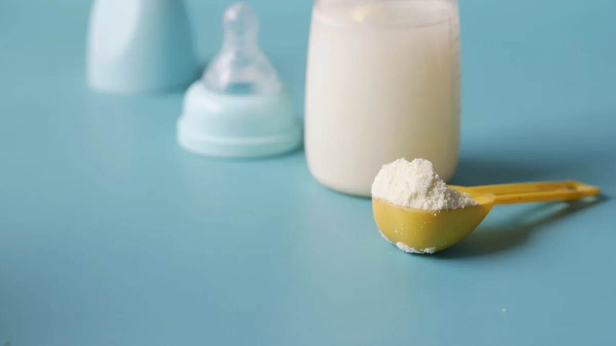 Baby bottle of milk and a scoop of formula powder beside a spare teat on a pale blue surface