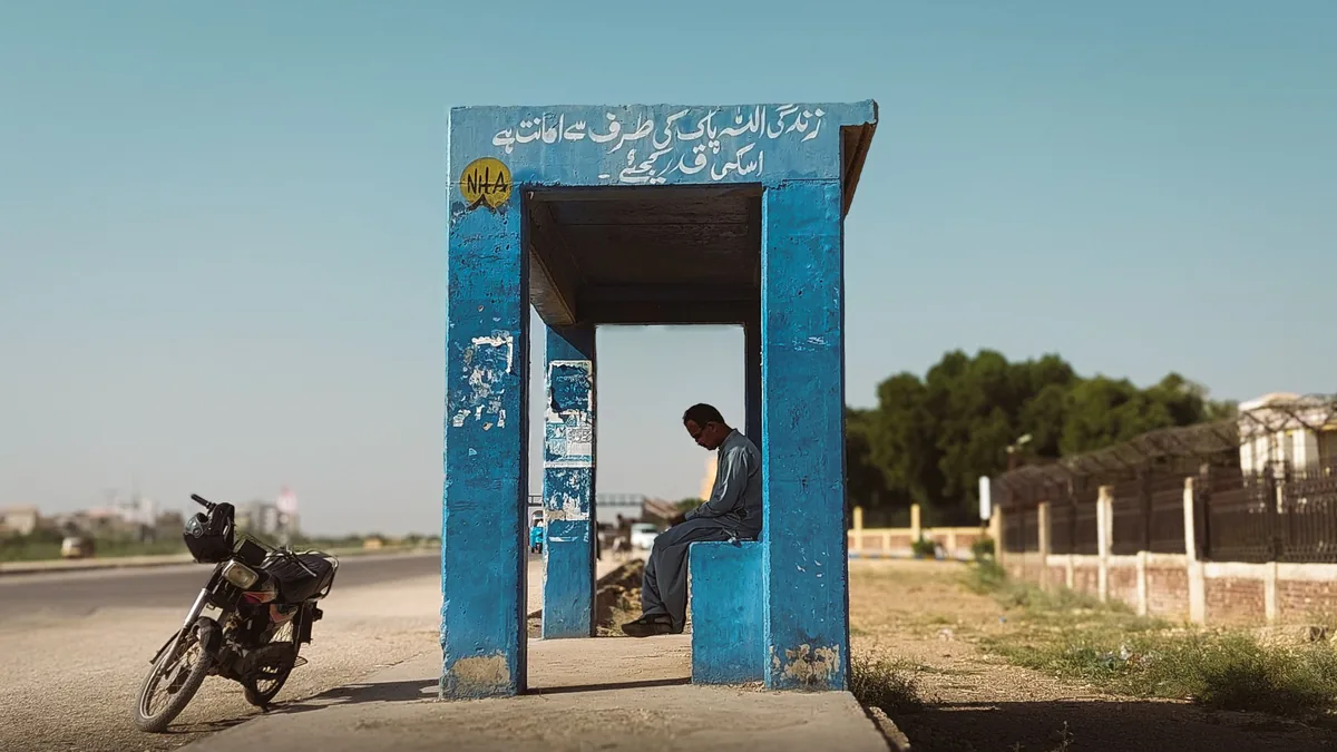 A blue roadside bus shelter with Urdu lettering beside a road in Pakistan, a man seated inside and a motorbike parked by it