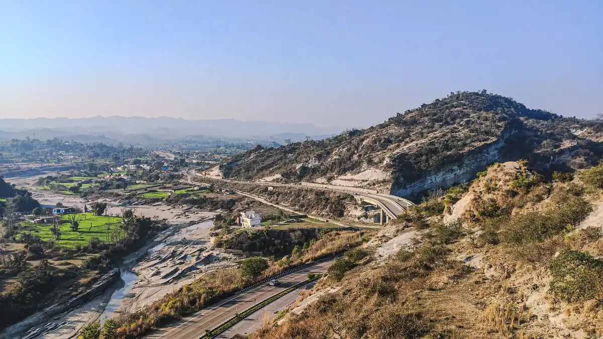 Dual carriageway curving through dry scrub-covered hills above a green river valley and town under a clear sky