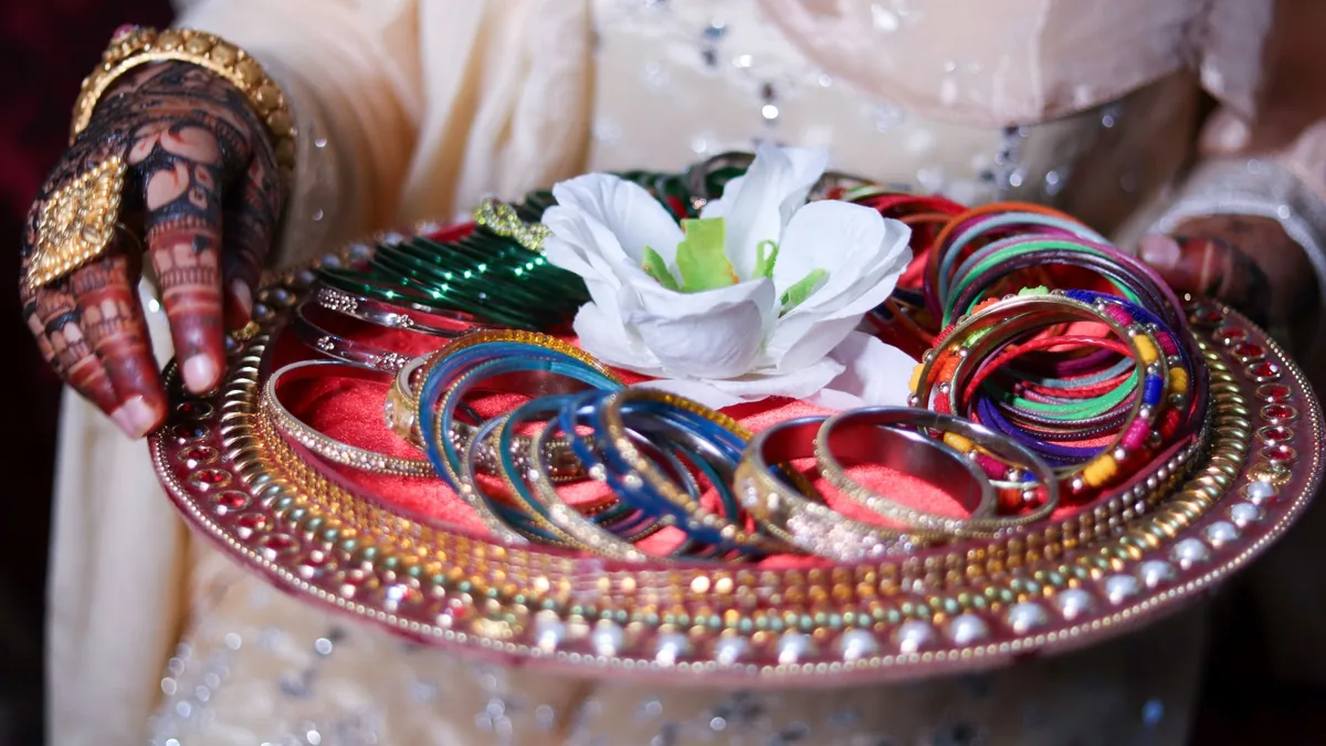 Henna-decorated hands holding a wedding tray of glass bangles with a white flower, in bridal clothing