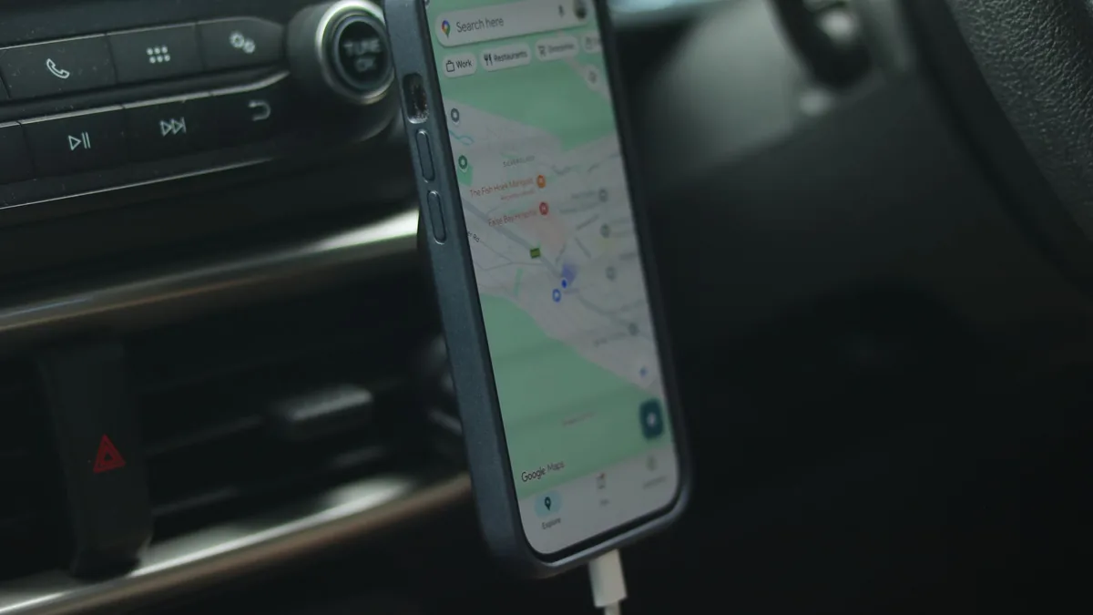 Smartphone in a dashboard mount showing a map navigation app inside a car, with a charging cable attached