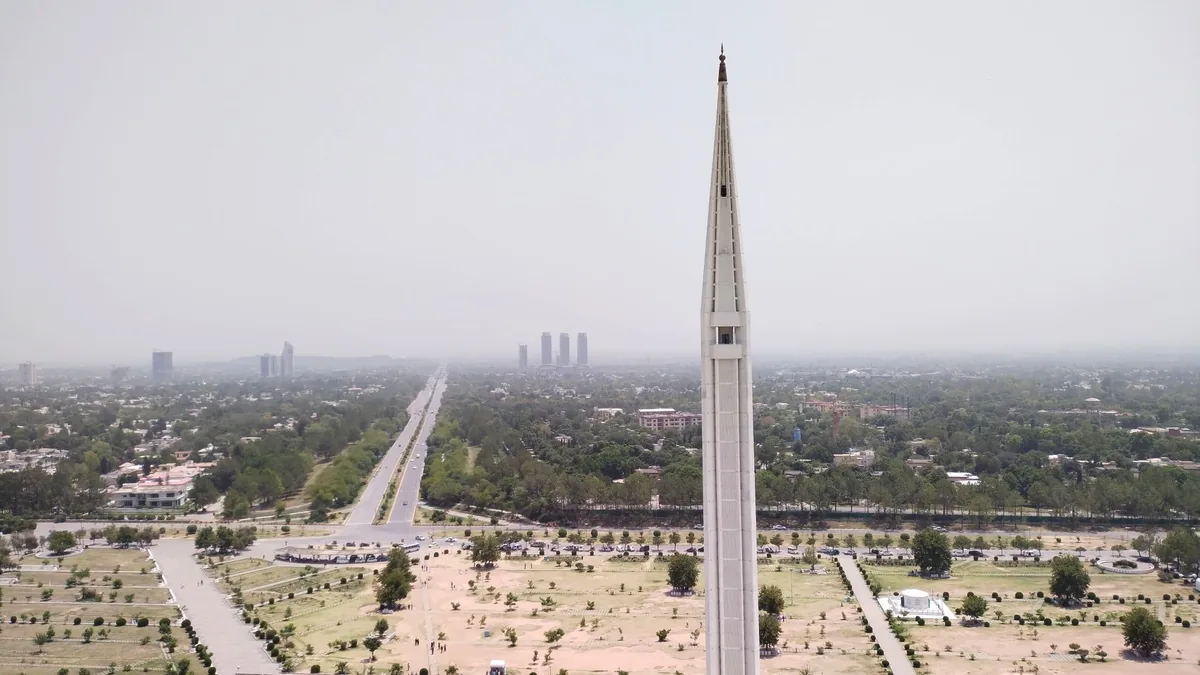 Aerial view past a Faisal Mosque minaret over landscaped grounds, a straight avenue and the hazy Islamabad skyline