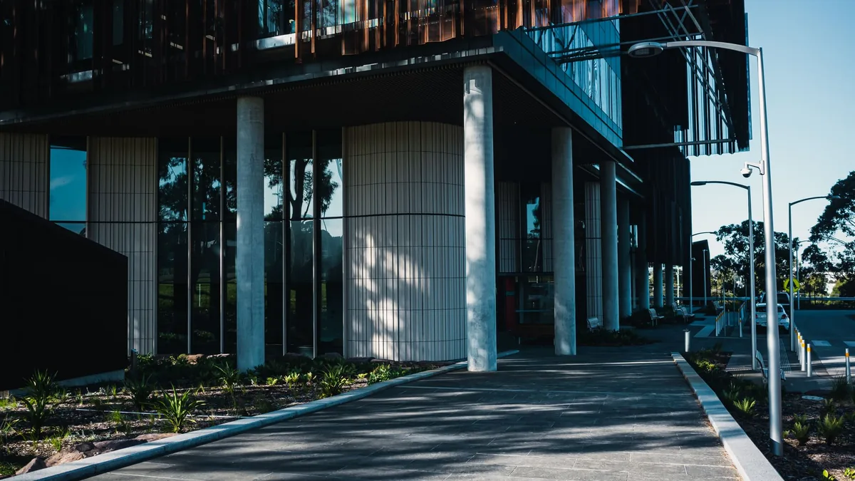 Modern hospital building with glass and timber facade, landscaped beds and a paved walkway leading to the entrance on a sunny day