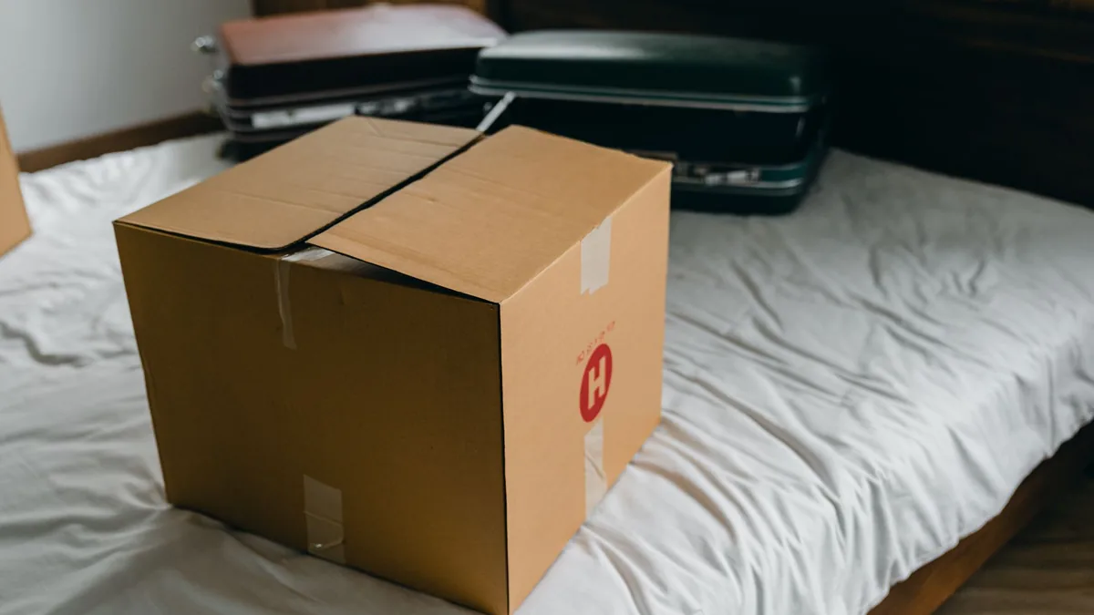 Taped cardboard moving box on a bare mattress with dark suitcases behind, belongings packed for a room move