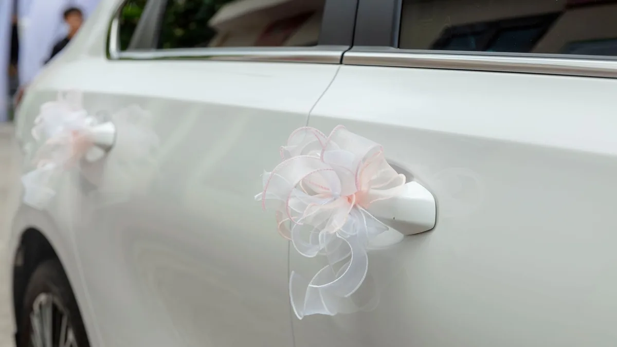 Close-up of a white ribbon bow tied around the door handle of a white car decorated for a wedding