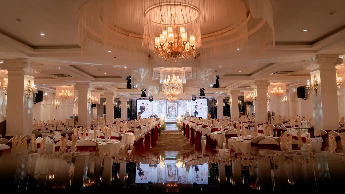 Empty banquet hall set for a wedding reception, rows of dressed tables under chandeliers and a lit stage at the far end