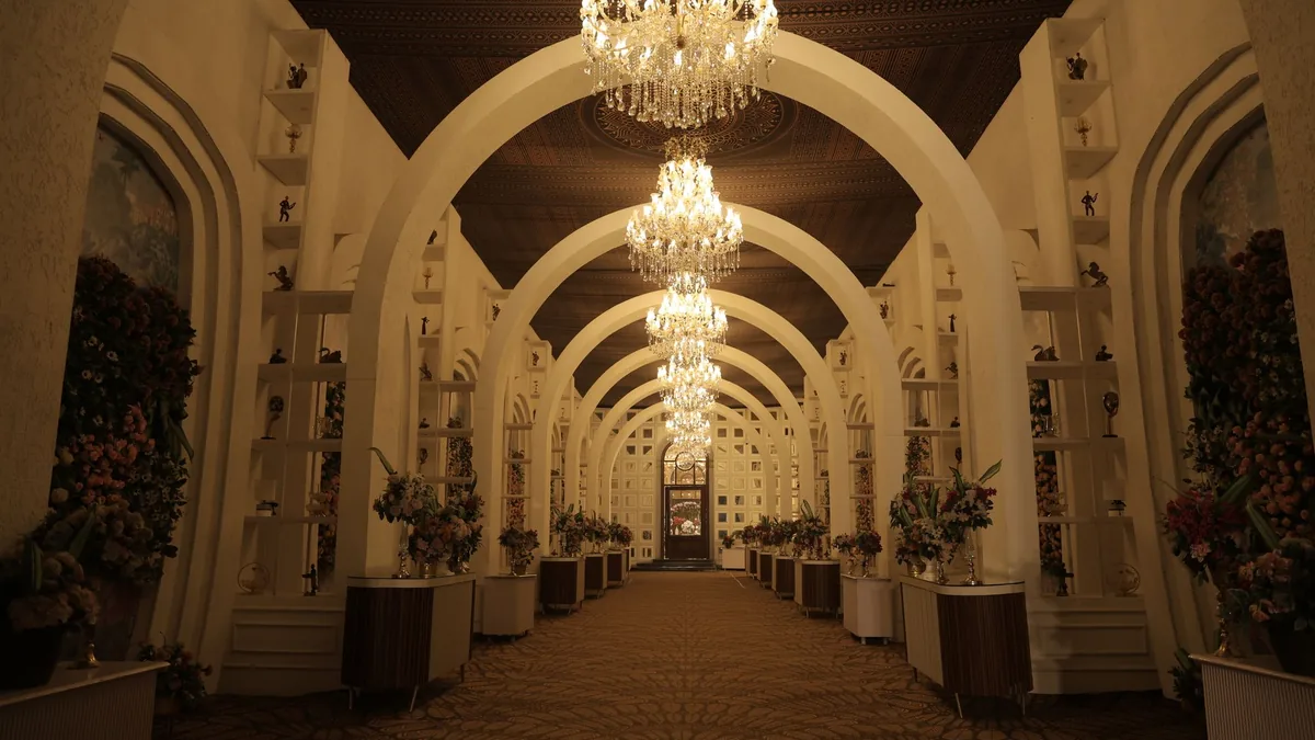 Empty arched corridor of a wedding venue lit by crystal chandeliers, lined with floral arrangements and a carpeted walkway
