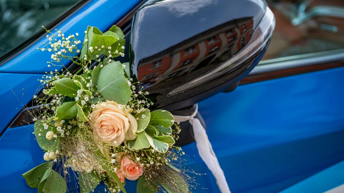 Close-up of a floral wedding decoration with peach roses and greenery tied beside the side mirror of a blue car