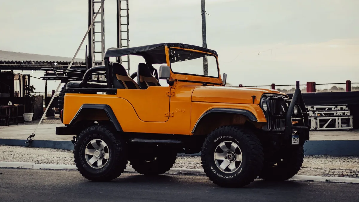 Orange classic open-top jeep with a black roll bar parked on a paved street under an overcast sky