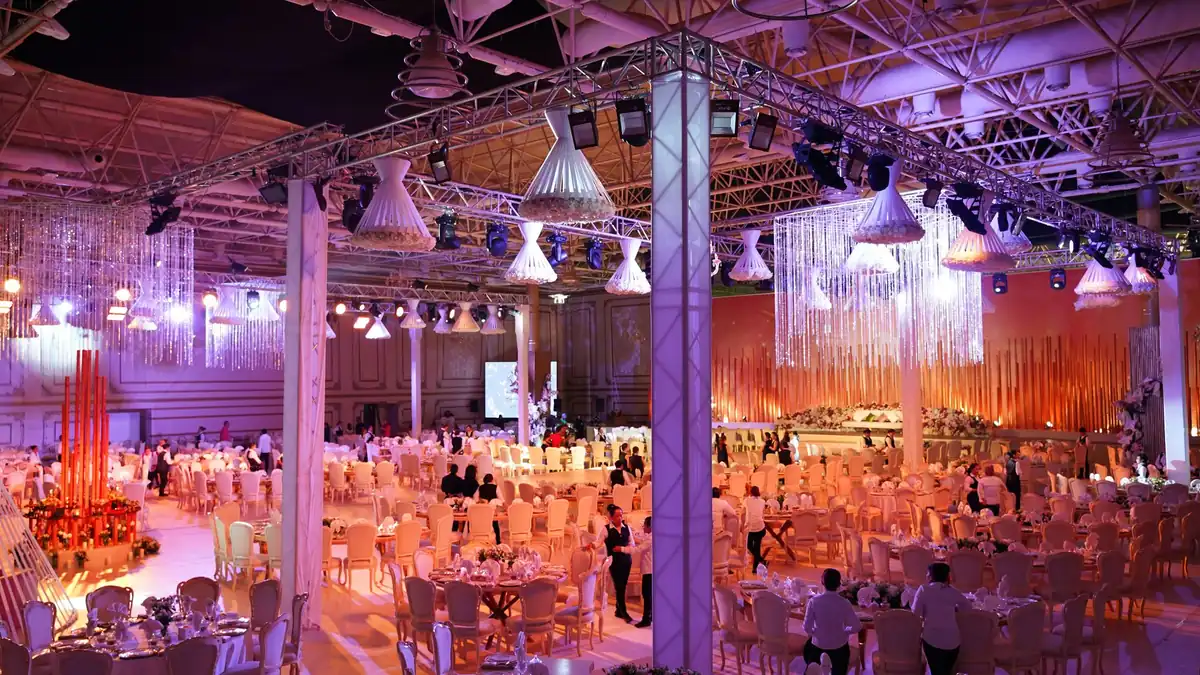 Large wedding hall under a steel roof with round dining tables, coloured lighting and guests moving between tables