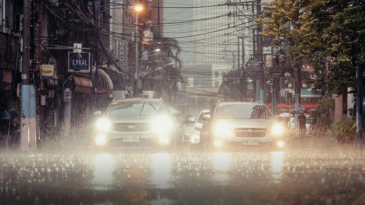 Cars with headlights on driving slowly along a city street in heavy rain, with wet road spray and overhead wires