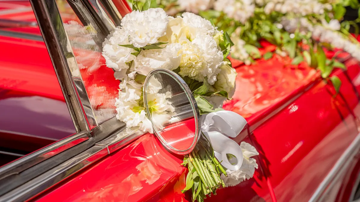 Close-up of a red car's side mirror and door decorated with a bouquet of white flowers and ribbon for a wedding