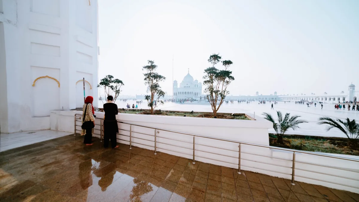 Two visitors seen from behind on a white marble terrace overlooking a wide courtyard with a domed gurdwara in the hazy distance