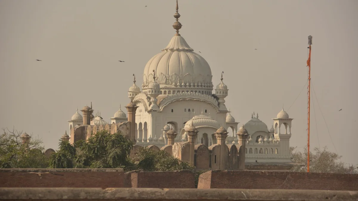 White domed Sikh-era mausoleum with small cupolas and a tall flagpole, seen over a brick wall in hazy light in Lahore