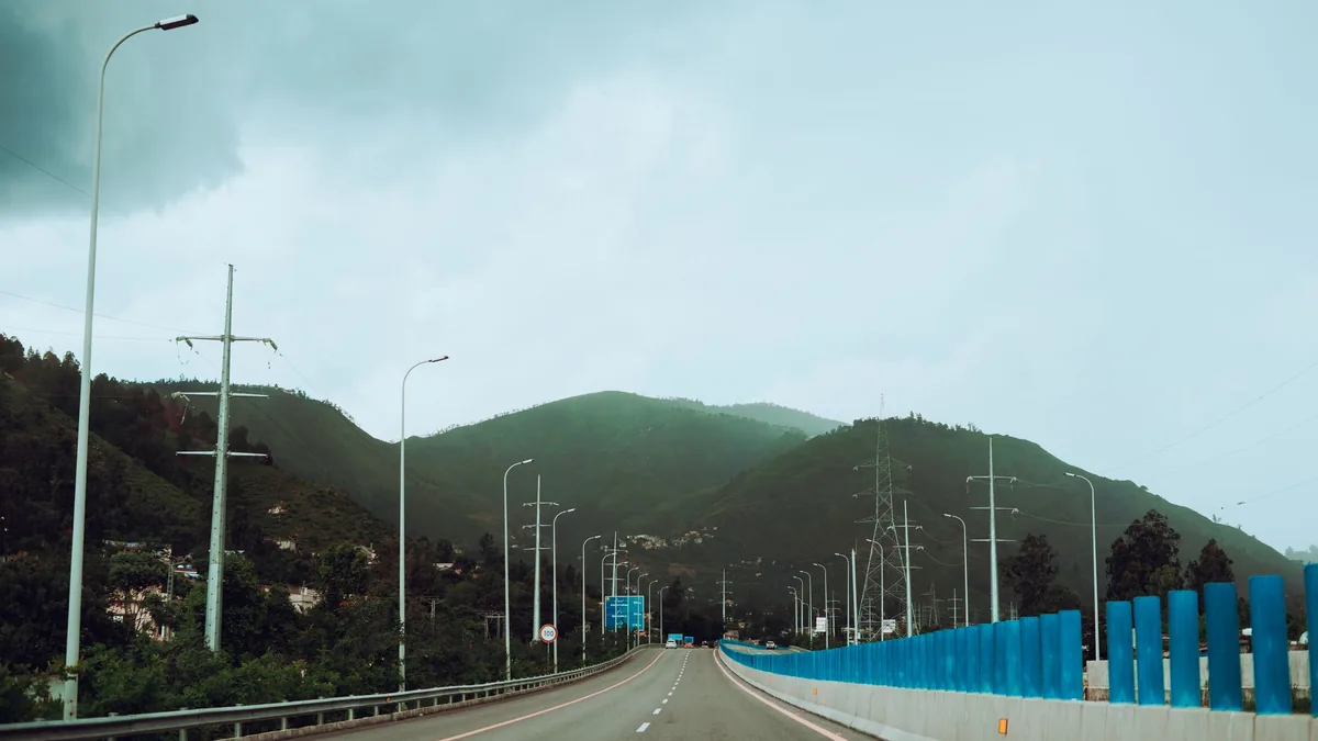 Quiet dual carriageway with street lights and a blue roadside barrier leading towards green hills under an overcast sky