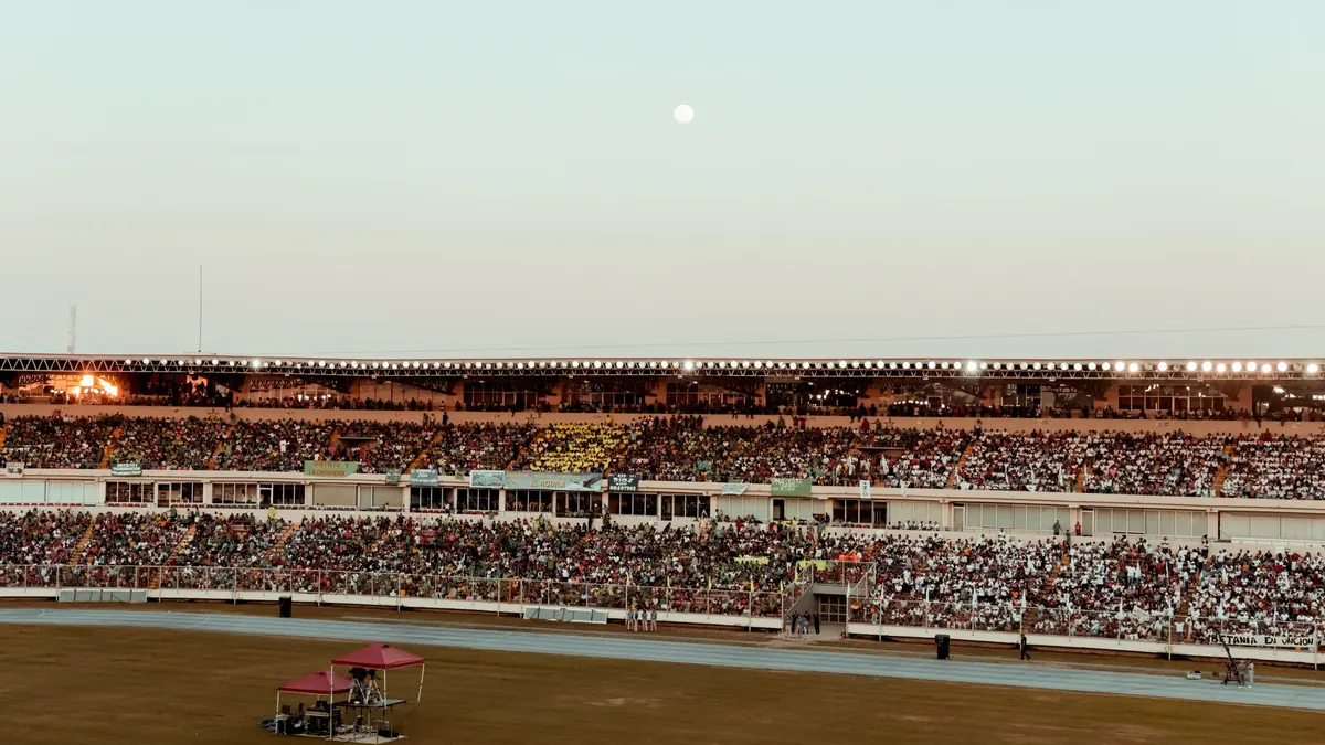 Packed stadium stands at dusk seen from across the ground, with a small covered cart parked on the field edge