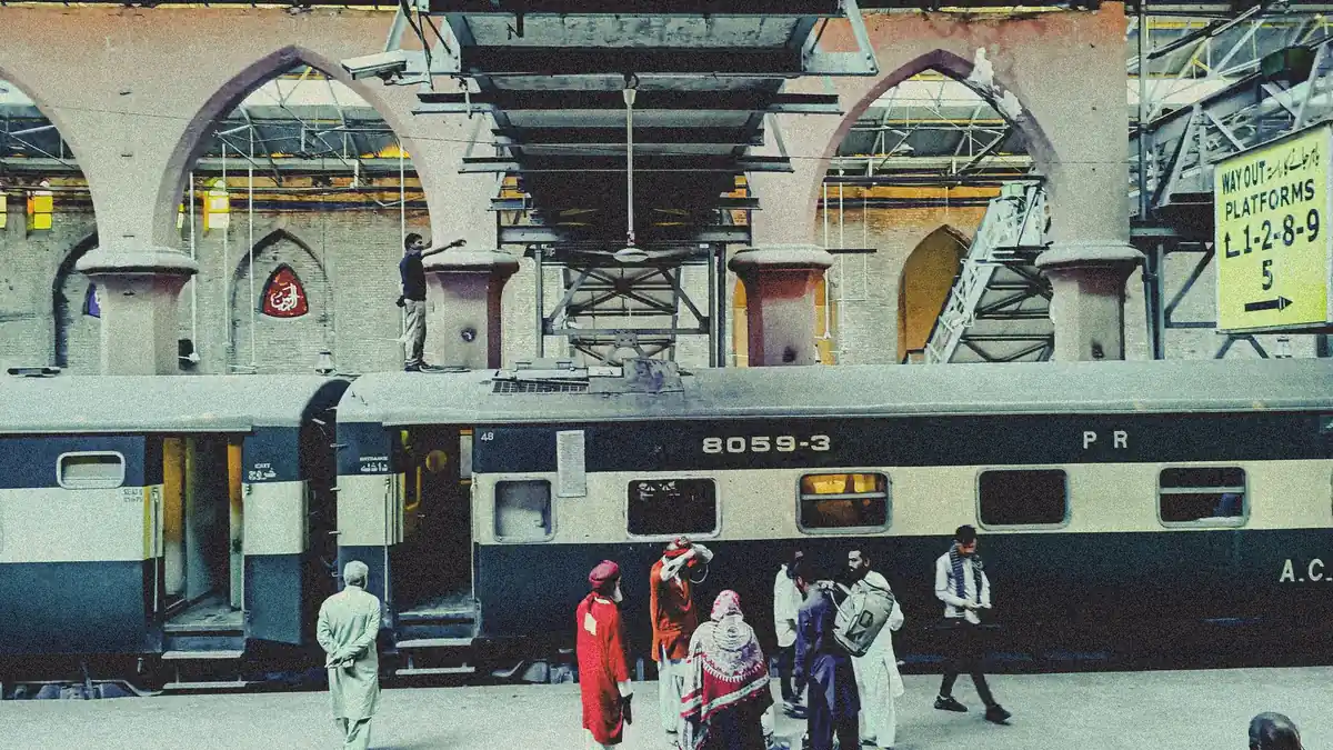 Passengers with bags beside a green and cream Pakistan Railways coach on an arched platform at Lahore Railway Station