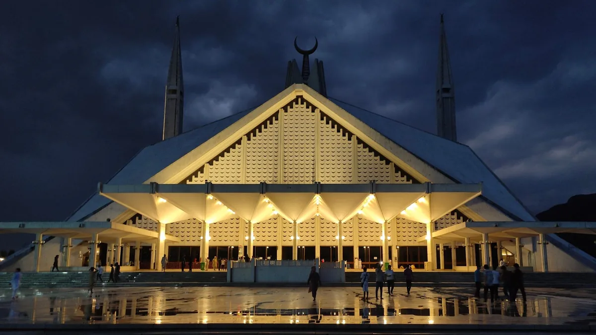Faisal Mosque in Islamabad lit up at dusk under a dark cloudy sky, its wet marble courtyard reflecting the lights