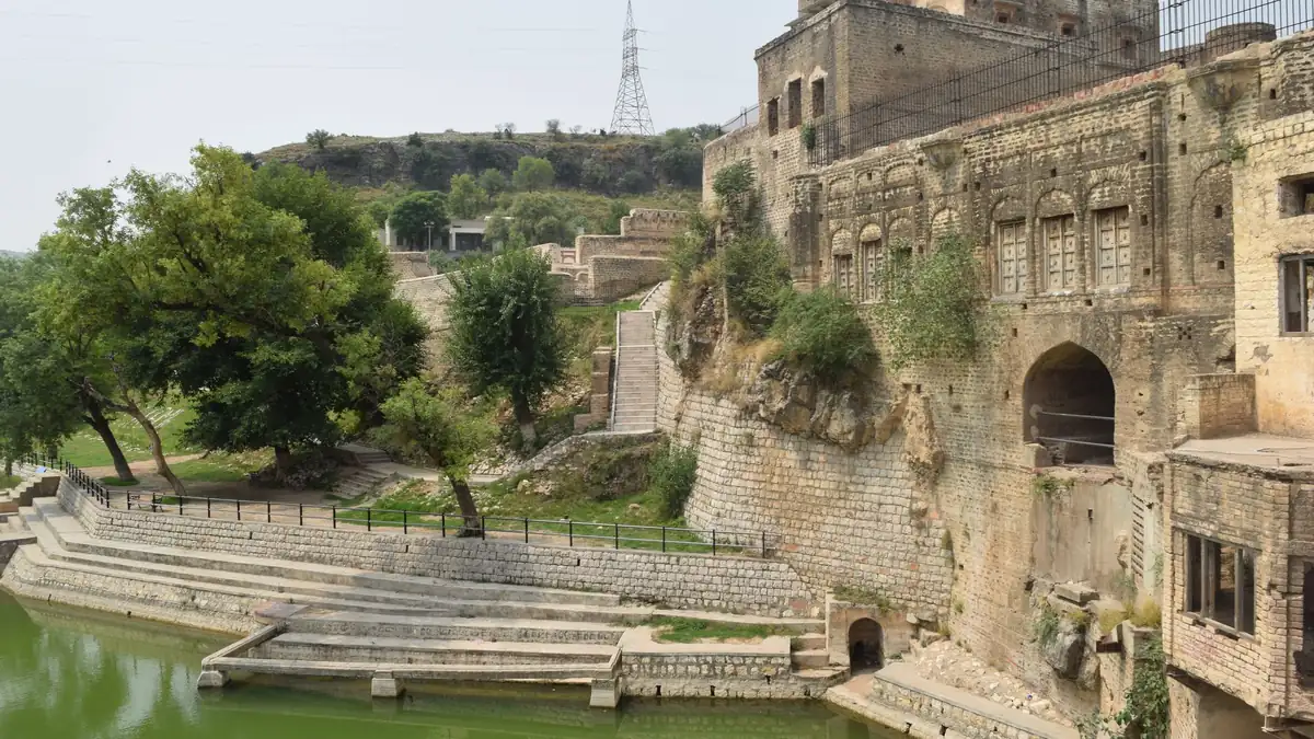 Green sacred pond with stone steps beside weathered old temple and haveli buildings at Katas Raj under a hazy sky