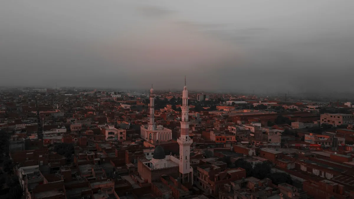 Aerial view of dense Gujranwala rooftops at dusk with twin mosque minarets rising under a hazy grey sky