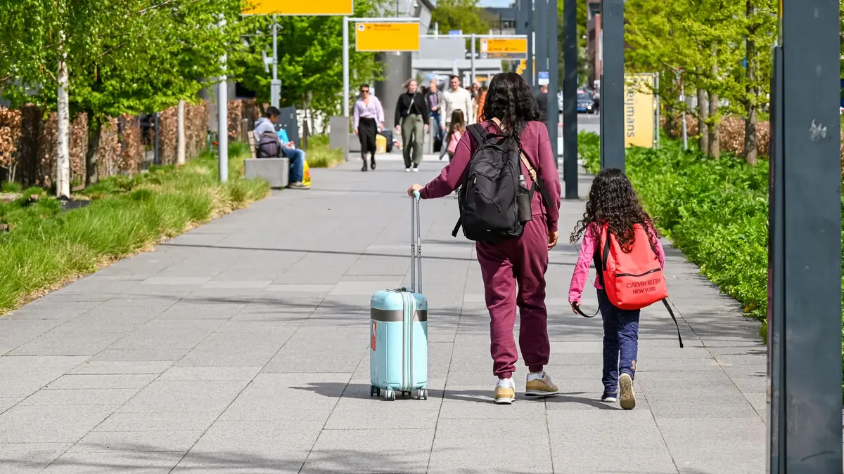 A mother and young daughter seen from behind, walking along a tree-lined airport path with a suitcase and backpacks