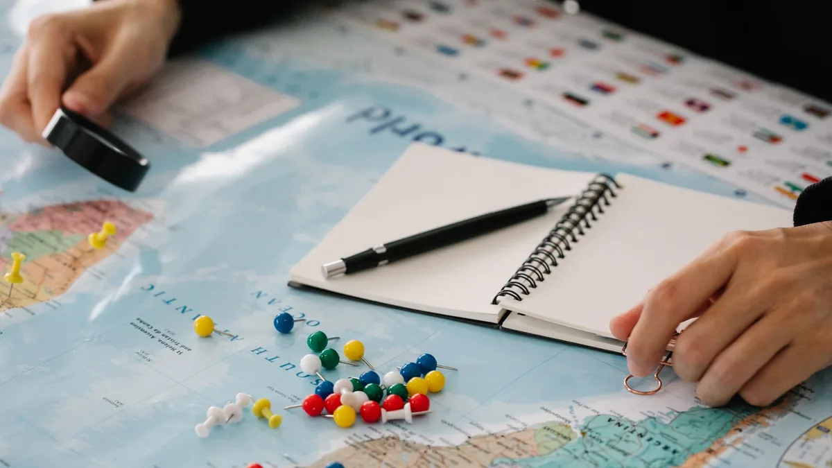 Hands planning a day route with a pen and spiral notebook beside a map dotted with coloured pins