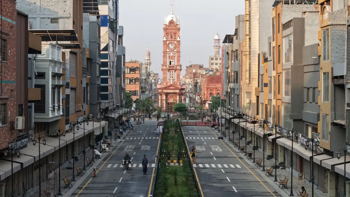 A wide bazaar road lined with shopfronts and a planted median, leading to the red Clock Tower at the far end